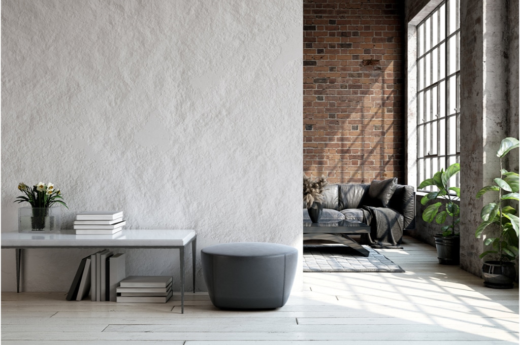 salon de type industriel avec un canapé noir et un mur en brique dans le fond. un mur blanc devant avec une console avec des livres et un tabouret.