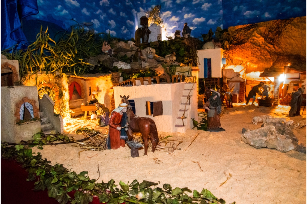 vue rapprochée sur une crèche avec des santons. on y voit des maisons dans du sable, des personnages, un cheval marron, le ciel est aussi représenté avec des nuages. des lumières émettent des petits éclairages lumineux de part et d'autres.