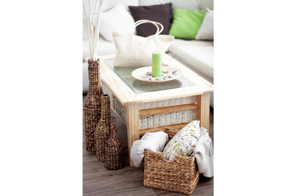 vue sur une table basse en bois naturel clair avec un plateau avec du verre au centre. des paniers et vases en osiers sont posés près de la table basse. une bougie dans une assiette est posée sur la table basse ainsi qu'un sac à main à anse. on aperçoit dans le fond de la photo, un canapé avec des coussins.